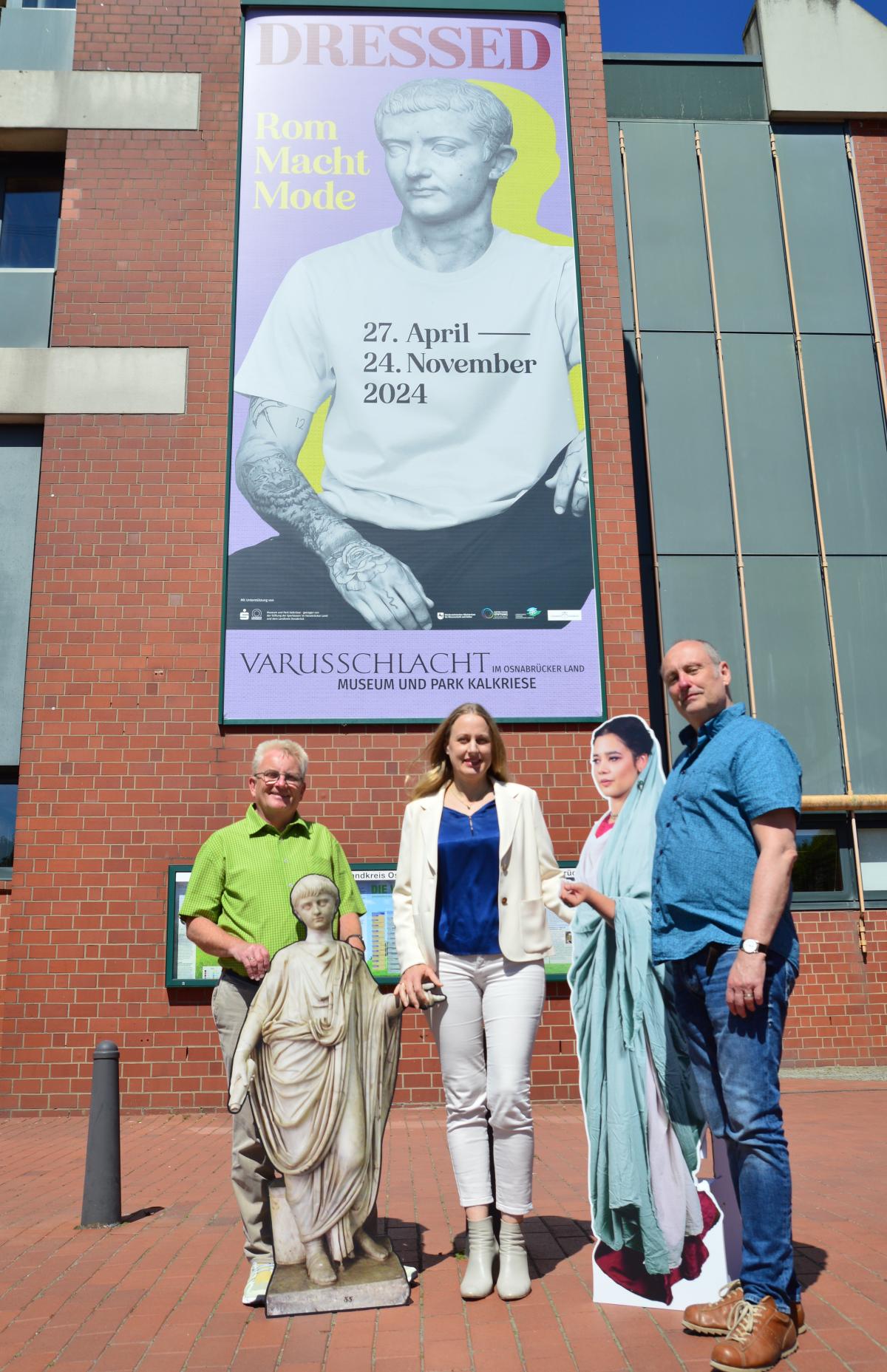 Drei Personen stehen vor einem großen Plakat an einer Hauswand.