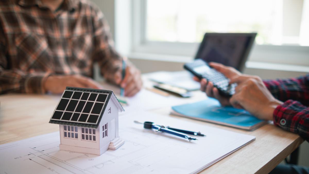 Zwei Personen sitzen an einem Schreibtisch, auf dem das Modell eines Hauses mit Solaranlage steht.