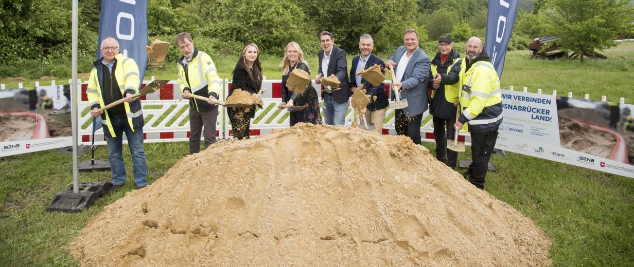 Eine Personengruppe mit Spaten in den Händen steht hinter einem Sandhaufen.