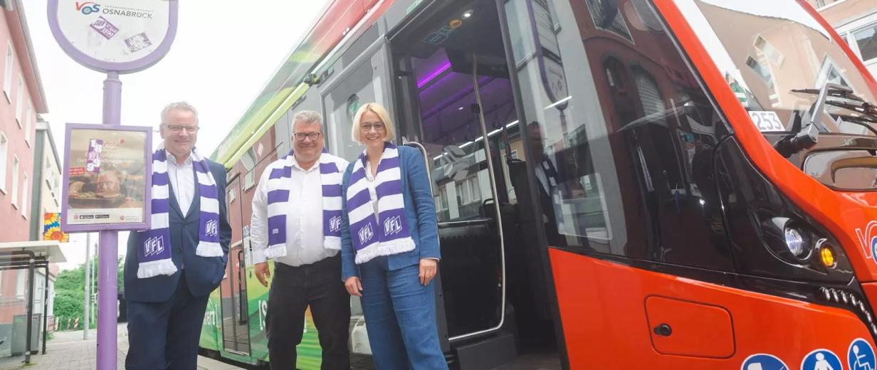 Drei Personen mit weiß-lila gestreiften Schals stehen vor einem Bus.