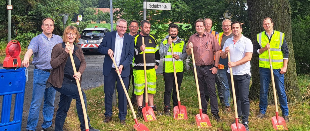 Eine Personengruppe mit schaufeln in den Händen steht an einer Straßenbaustelle.