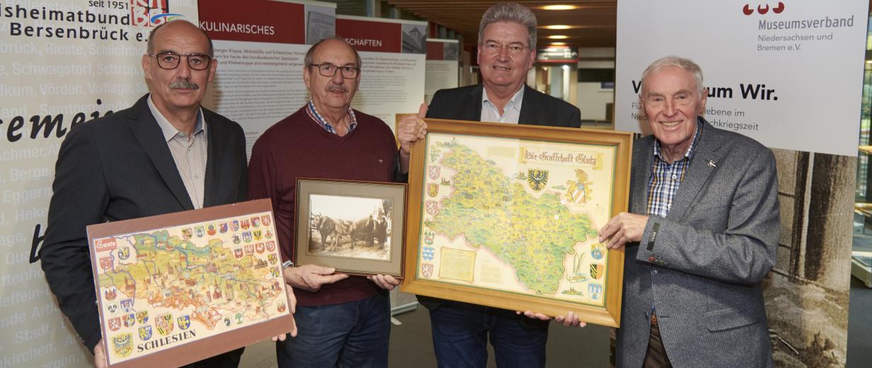 Vier Personen stehen vor den Postern einer Ausstellung und halten historische Karten in den Händen.