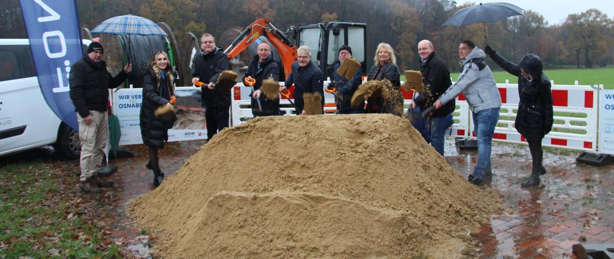 Eine Personengruppe mit Spaten in den Händen steht hinter einem Sandhaufen.