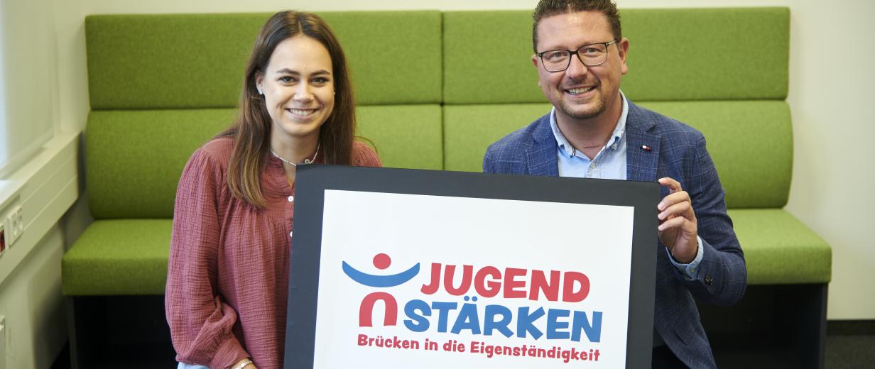 Zwei Personen mit einer Tafel in der Hand sitzen vor einer Bank.