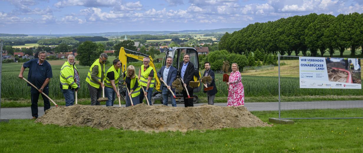 Eine Personengruppe mit Schaufeln in den Händen steht hinter einem Sandhaufen.