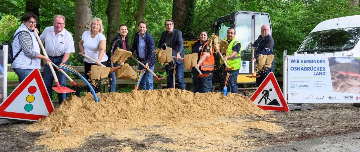 Eine Personengruppe mit Schaufeln steht hinter einem Sandhaufen.