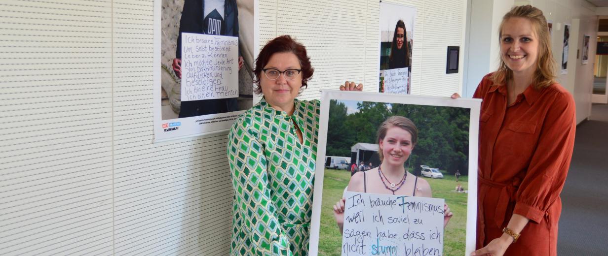 Zwei Personen mit einem Poster in den Händen stehen in einem Gebäudeflur.