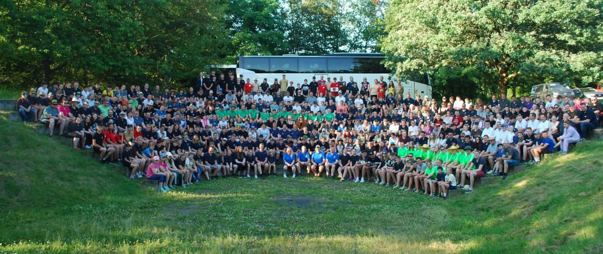 Eine große Personengruppe sitzt wie in einem Amphitheater auf einer Wiese.