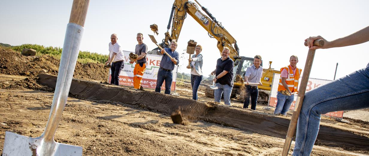 Eine Personengruppe mit Spaten in den Händen steht auf einer Baustelle.