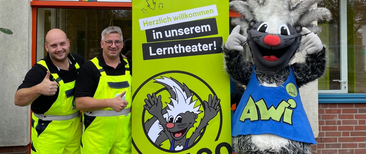 Zwei Personen und ein Maskottchen stehen neben einem Roll Up Poster.