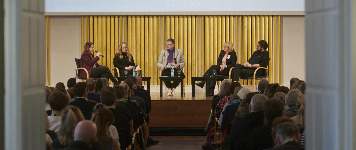 Fünf Personen sitzen auf einer Bühne in einem Saal vor Publikum.