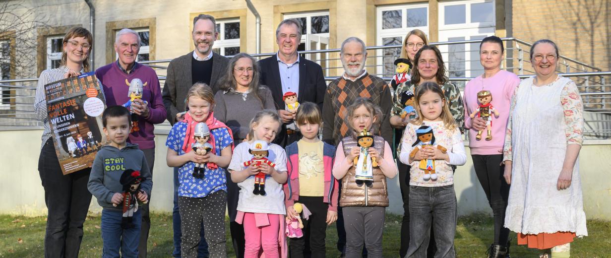 Eine Personengruppe mit Playmobilfiguren in den Händen steht vor einem Gebäude.