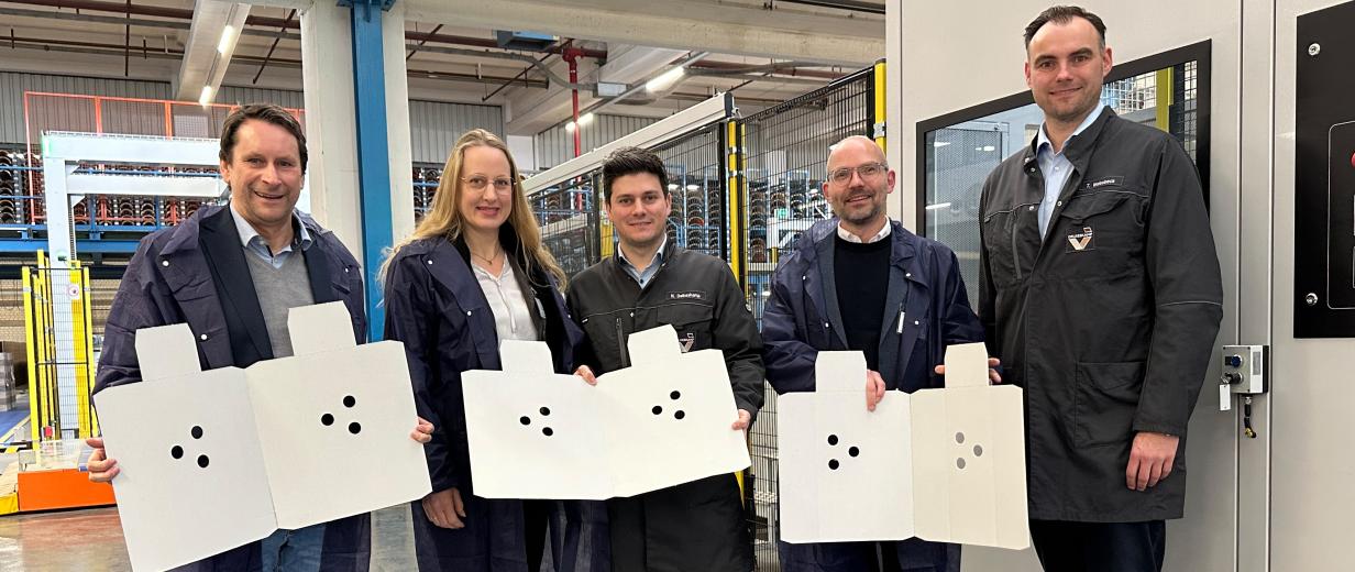 Fünf Personen mit Verpackungsmaterial in den Händen stehen in einer Werkhalle.