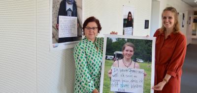 Zwei Personen mit einem Poster in den Händen stehen in einem Gebäudeflur.