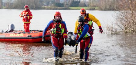 Zwei Personen in Schutzanzügen am Ufer eines Sees tragen eine Person auf einer Trage