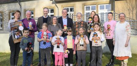 Eine Personengruppe mit Playmobilfiguren in den Händen steht vor einem Gebäude.