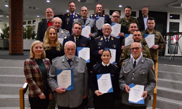 Eine Personengruppe steht in einem Gebäude auf einer Treppe. Einige Personen tragen Uniform des Bundeswehr und halten Urkunden in den Händen.