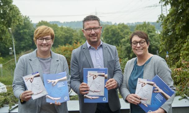 Drei Personen mit Broschüren in den Händen stehen auf einer Dachterrasse.