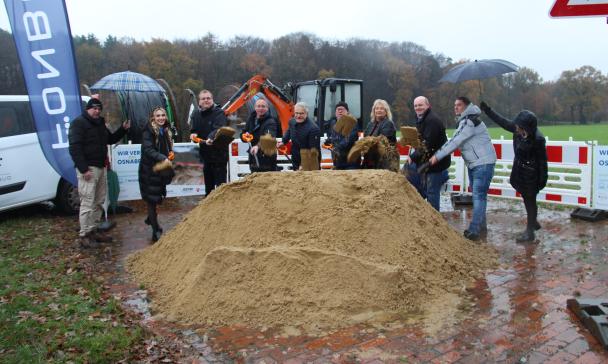 Eine Personengruppe mit Spaten in den Händen steht hinter einem Sandhaufen.