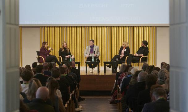 Fünf Personen sitzen auf einer Bühne in einem Saal vor Publikum.